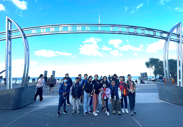 同學團隊於澳洲黃金海岸著名景點 Surfers Paradise 拍攝合照,背景為海灘與地標拱門 | 澳洲遊學團