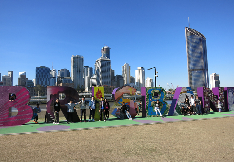 同學站在彩色 Brisbane 地標前拍照留念,背景為市中心高樓大廈與藍天,展現城市風貌 | 澳洲遊學團