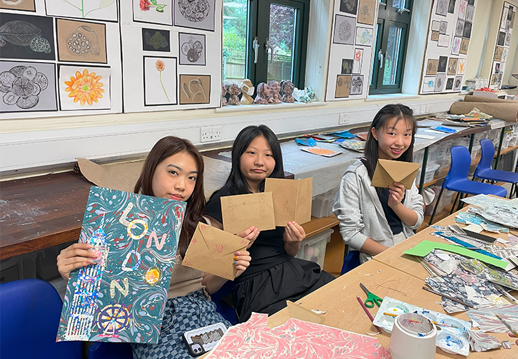 同學在美術教室中展示自己創作的信封與藝術作品,背景牆上掛滿素描畫作,展現學員的創意與課堂成果 | 英國遊學團