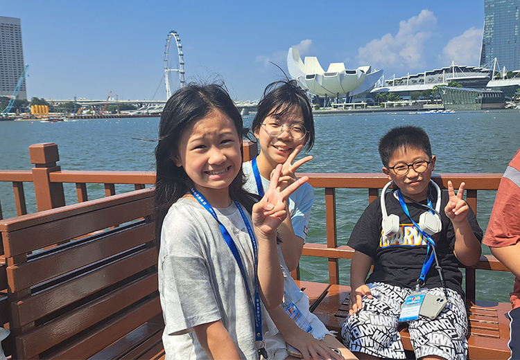 同學坐在濱海灣木製長椅上合影,遠眺摩天輪與藝術科學博物館建築 | 新加坡遊學團