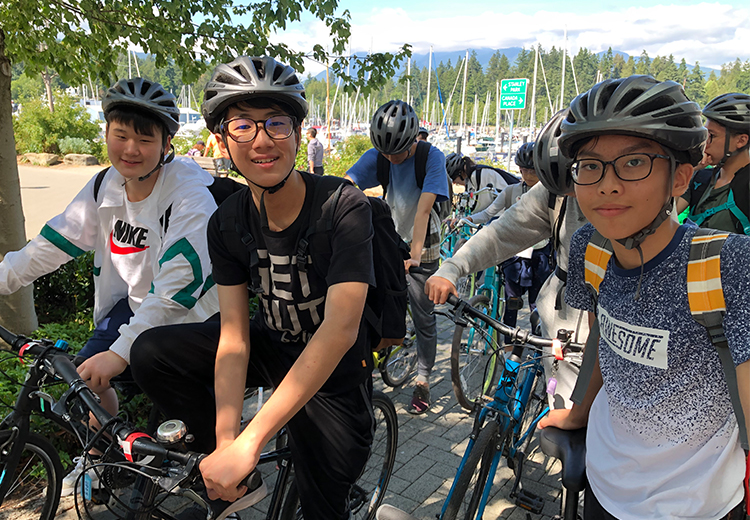 同學於溫哥華史丹利公園海邊騎單車,頭戴安全帽展現活力青春,背景可見遊艇與遠山景致 | 加拿大遊學團